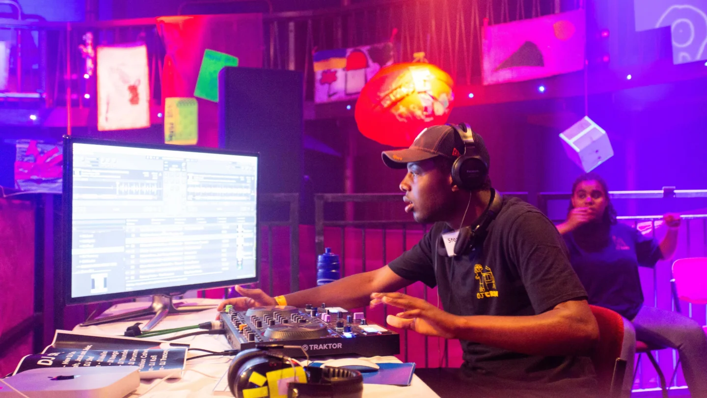A photo of a young black man wearing a black t-shirt and cap at a DJ deck.