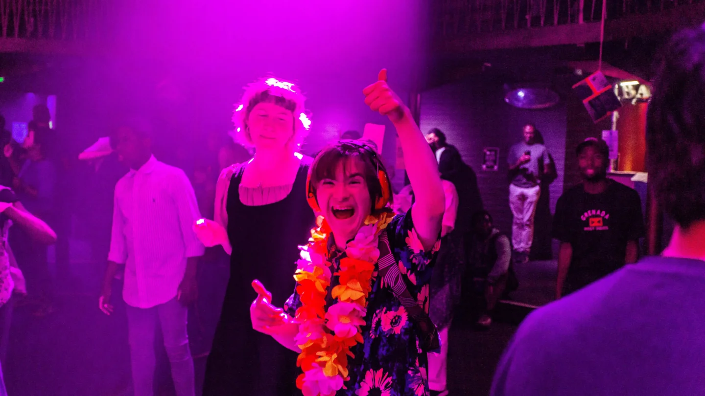 A photo of a young person wearing a floral top and hawaiian floral garland giving a thumbs up. They are on a dancefloor and bathed in pink light.