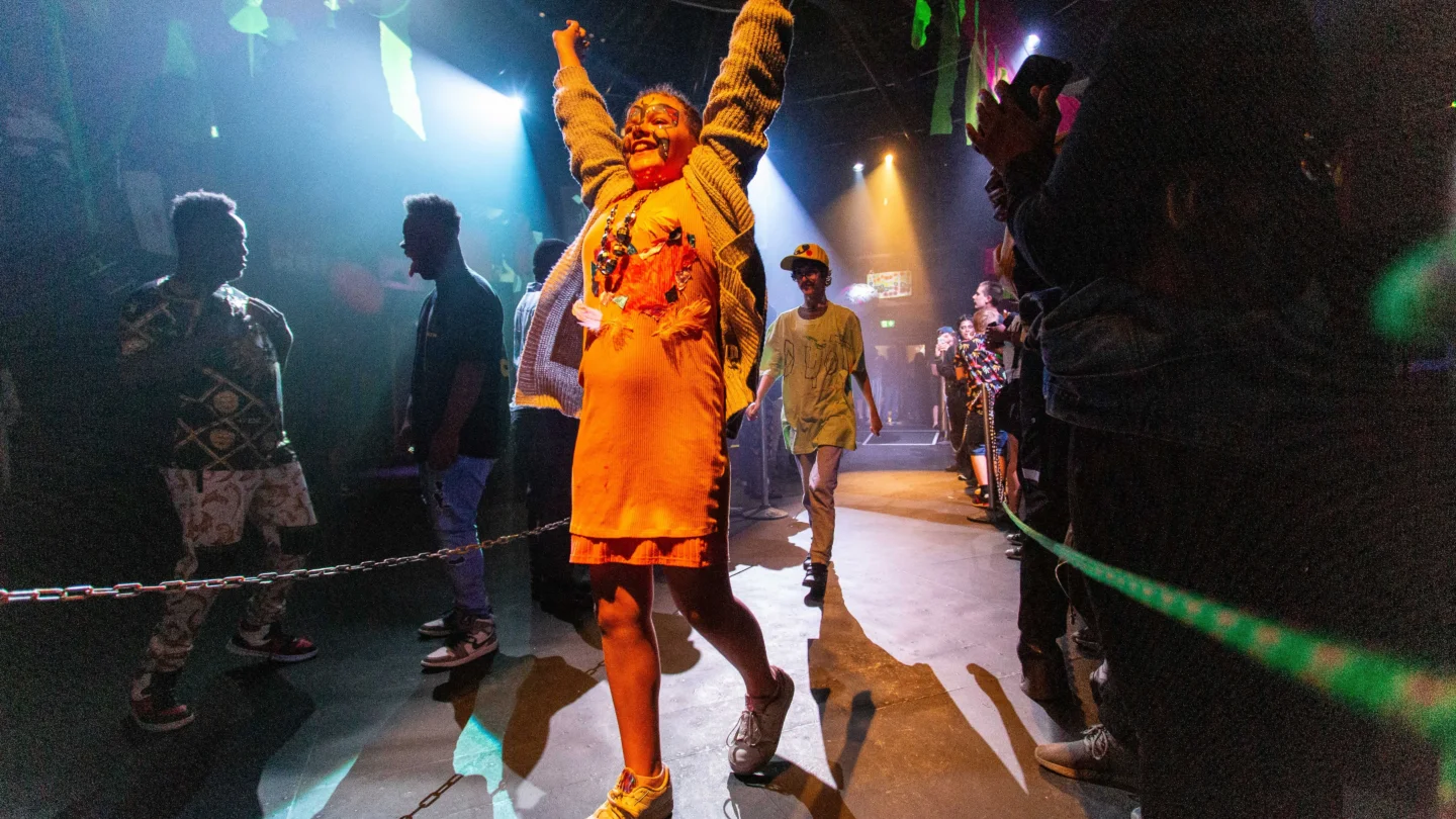 A photo of a young girl wearing orange on a catwalk, with her arms raised in celebration above her head.