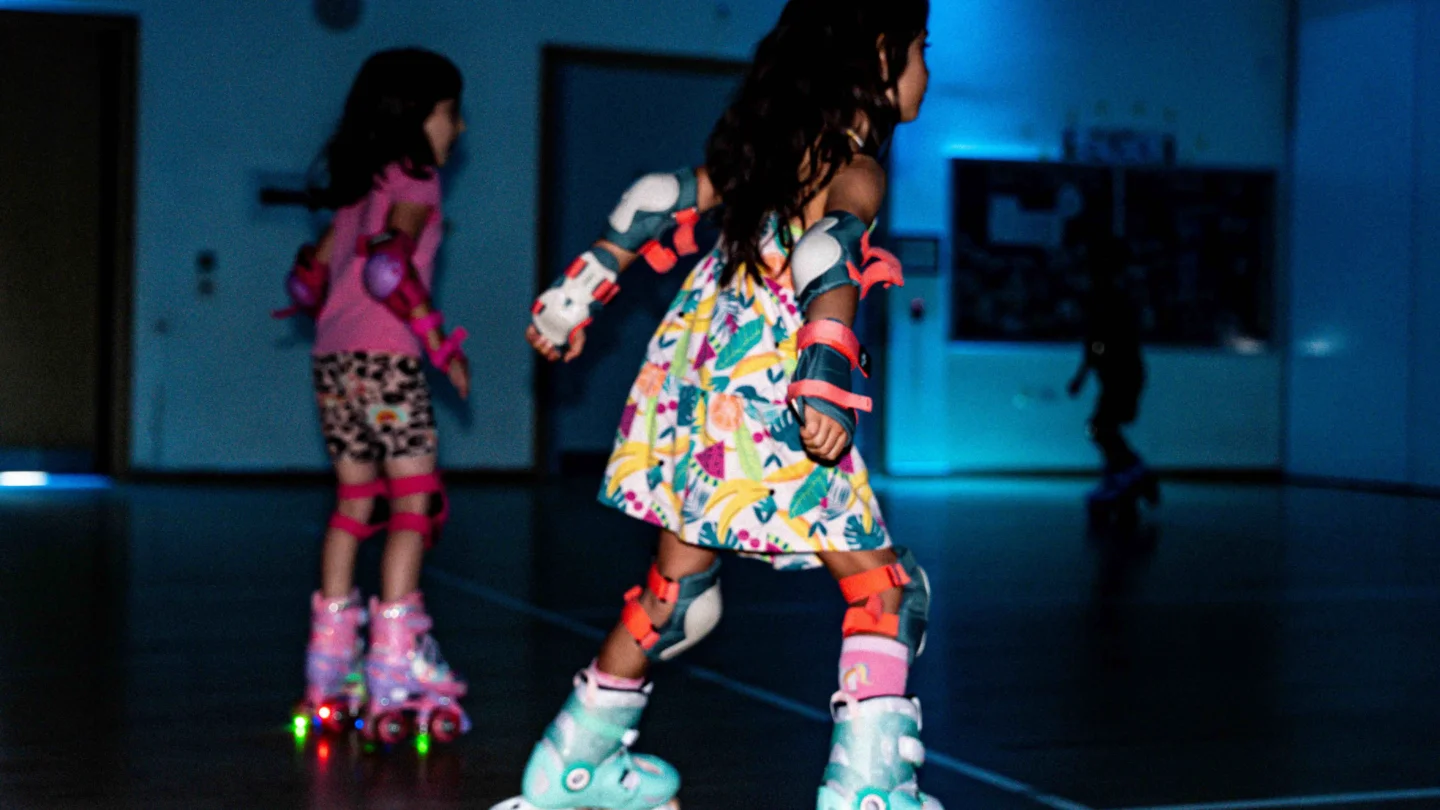 A photo of two young girls wearing brightly coloured outfits and roller skates, skating round a hall