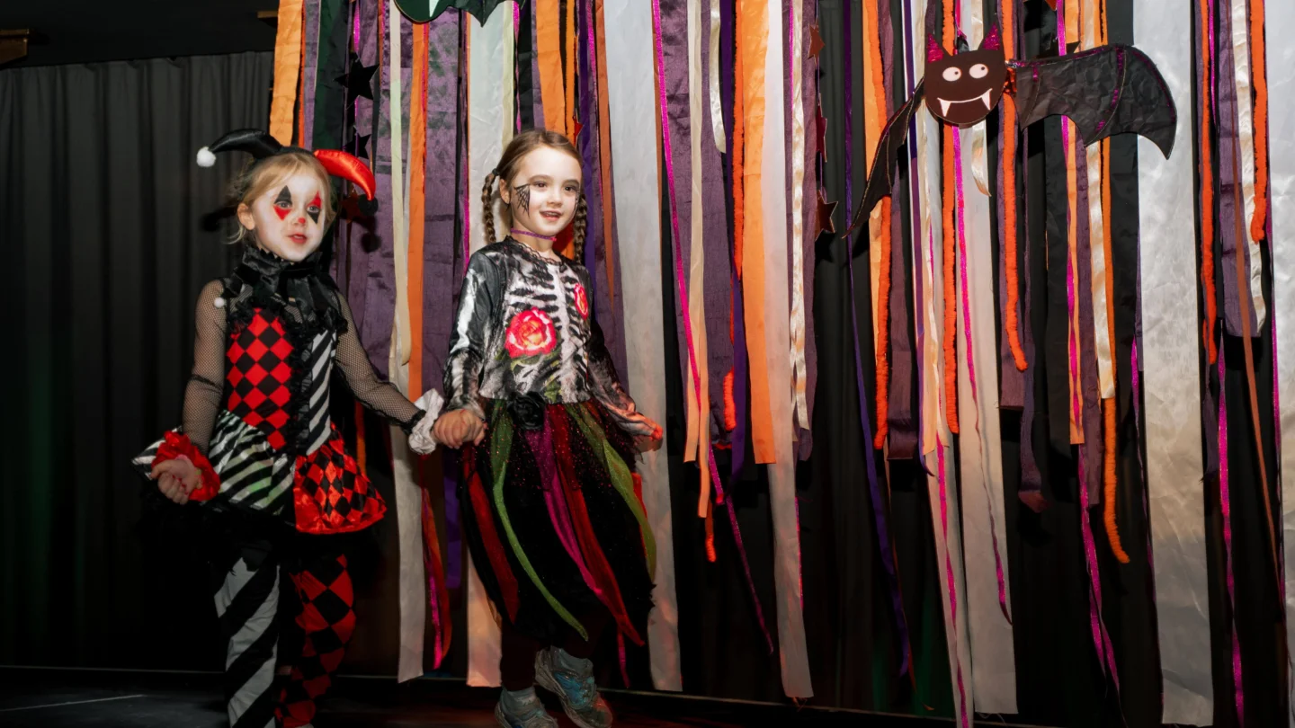 A photo of two children in fancy dress walking in front of a backdrop with black and orange streamers and hanging bats