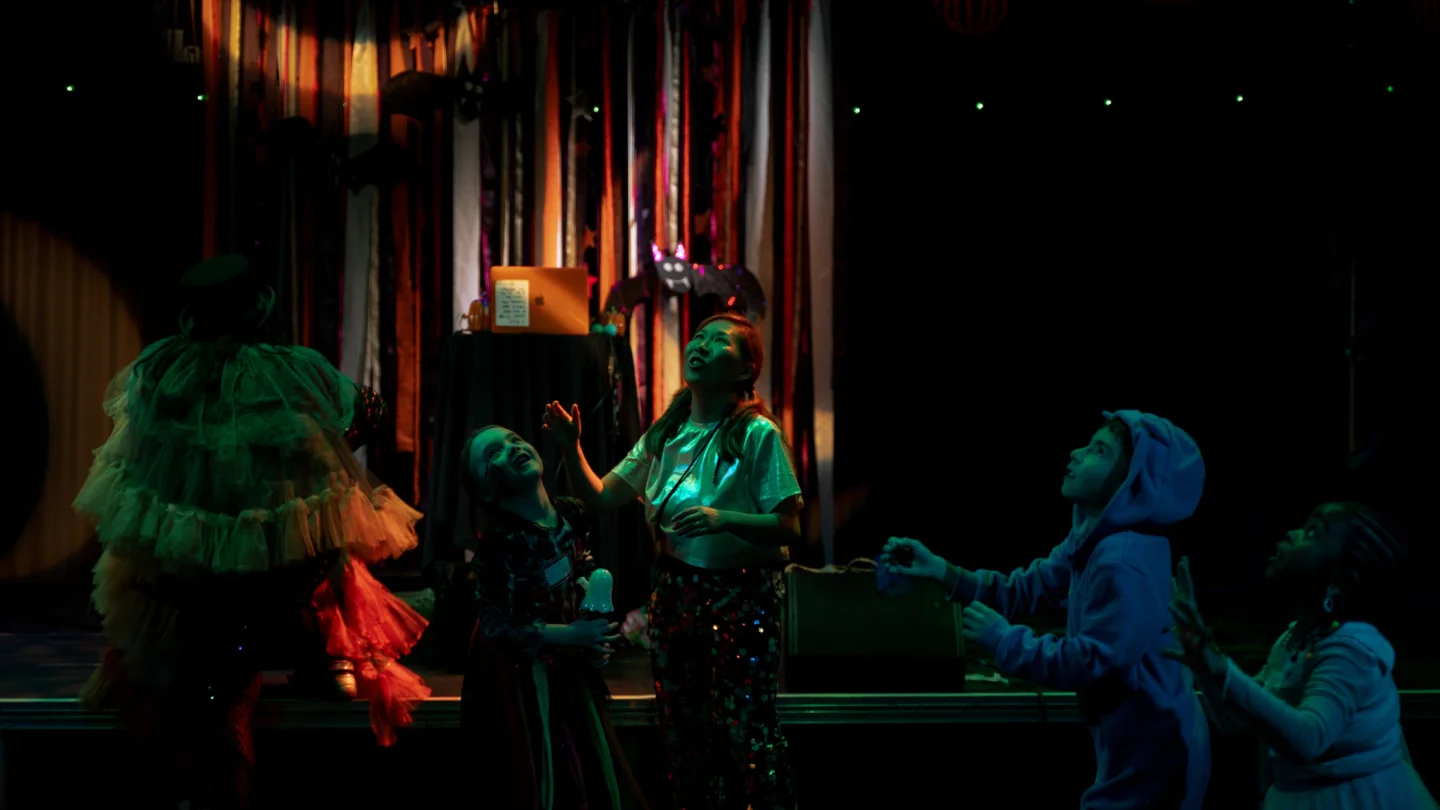 A photo of a dark dancefloor with orange and purple streamers along the back wall, and parents and children dancing.