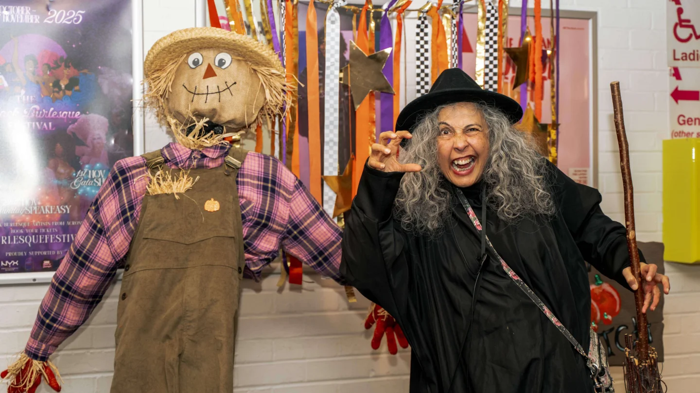 A photo of a woman with grey curly hair wearing a witch costume posing next to a scarecrow.