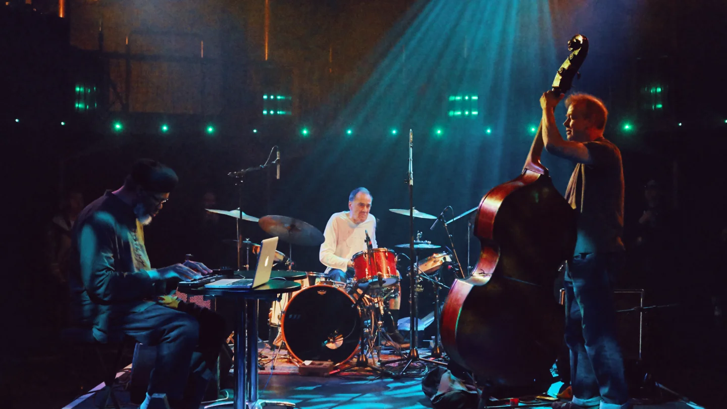 A photo of a band on stage under a blue lighting wash. There is a drumkit, keyboard and double bass.