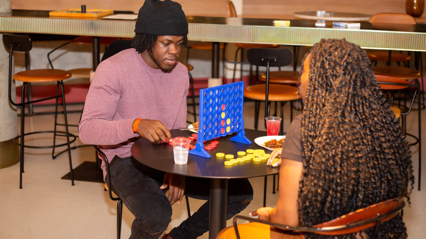 Two people, a black man wearing a purple jumper and beanie, and a woman with long braids wearing yellow trousers, sit at a table playing connect4.