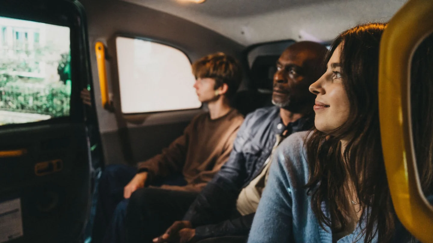 A photo of three people - a white woman with long brown hair, black man and young white man, in the back of a black cab.