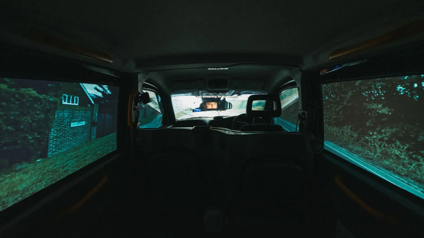 A photo of the interior of Driving Home - an installation inside a black cab with projection on the front windscreen and side windows