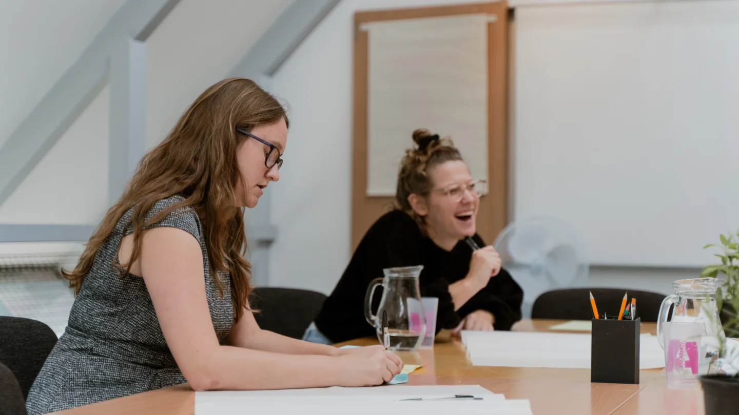 A photo of two white women in a meeting room. One is concentrating on something she is writing, and one is laughing during a conversation with someone out of shot. On the table there are pens, paper and jugs of water.