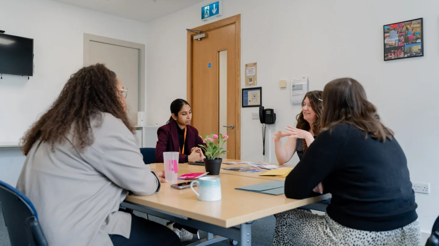 A group of women sit around a square table having a conversation. On the table there are mugs, workbooks and a pot plant.