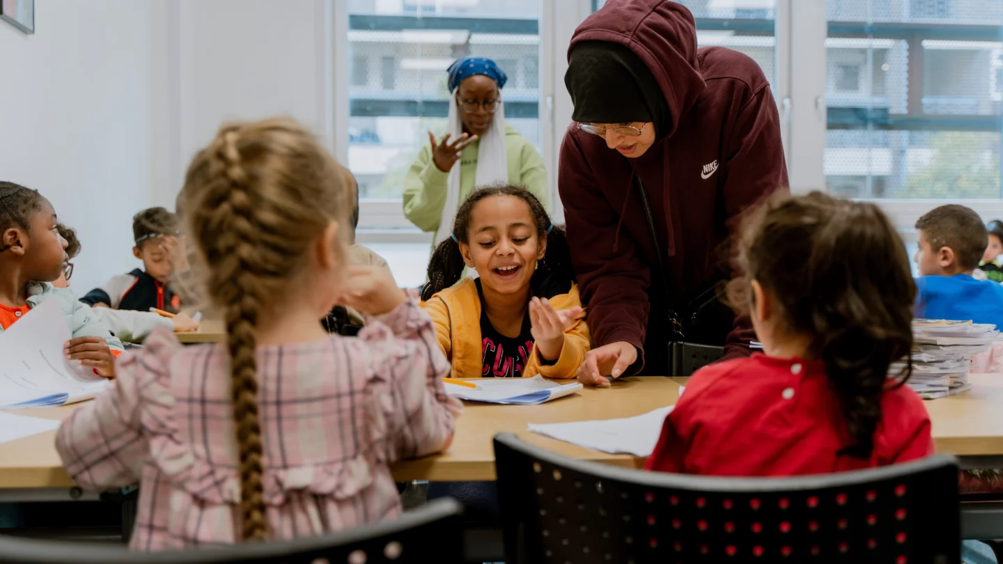 A photo of a lesson for children. A woman wearing a red hoodie is leaning over a young girl and pointing to something in her workbook. Other children are sat around working and chatting.