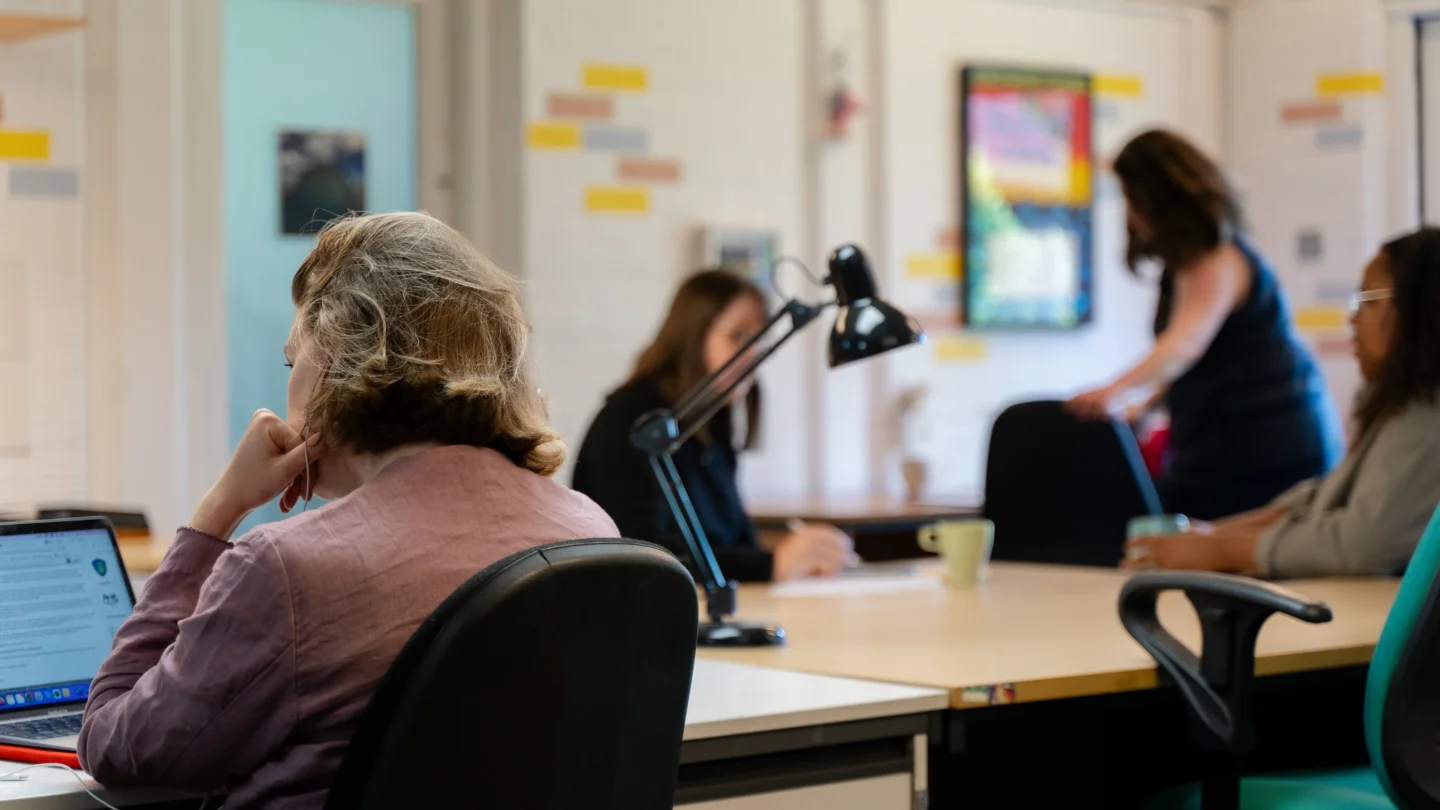 A shot from behind of a woman with short brown hair wearing a pink shirt. She is sat at a desk looking at a laptop. In the background there are other people working.