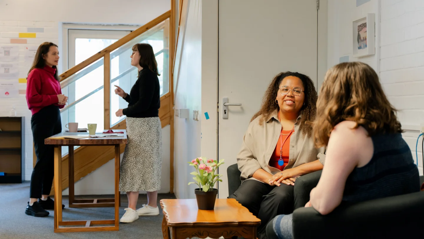 Shows an office with two people sat at a desk chatting in the foreground and two people standing in discussion in the background