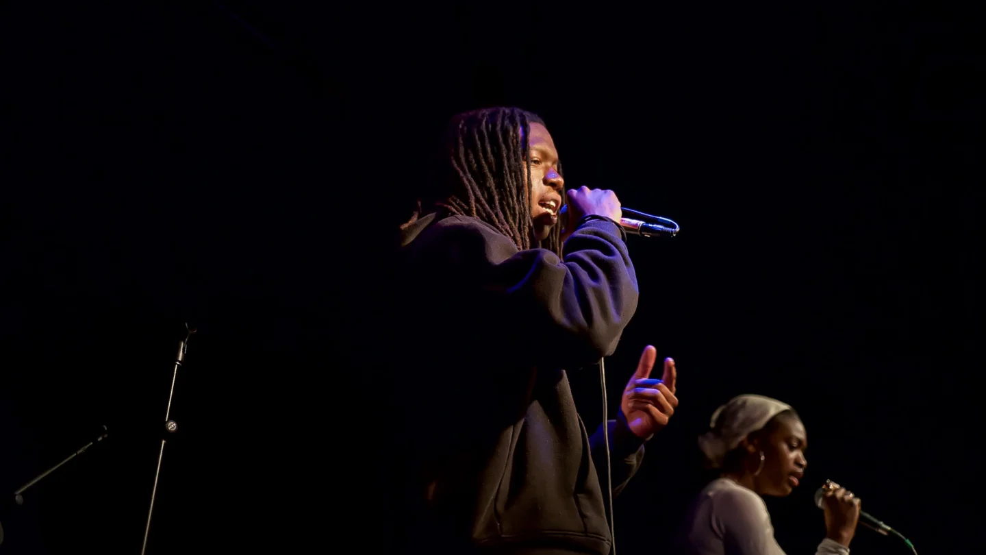 A young man with dreadlocks sings energetically into a microphone, immersed in his music. There is a young female performer in the background also singing into a microphone.