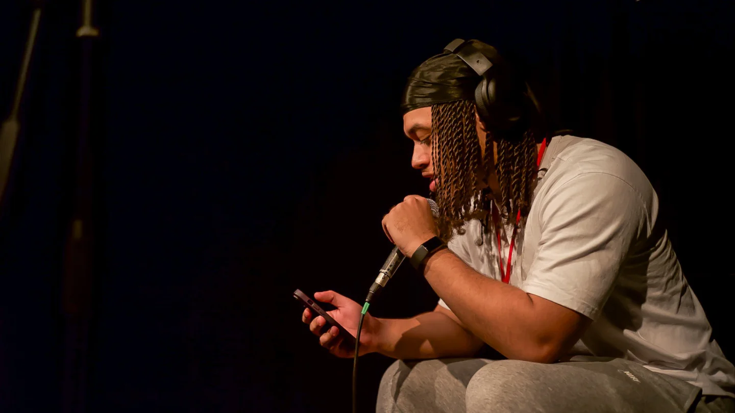 A young man, lit by a spotlight and wearing a pair of headphones, is holding a microphone in one hand and their phone in the other. They are mid-performance and appear calm and oblivious of the camera.
