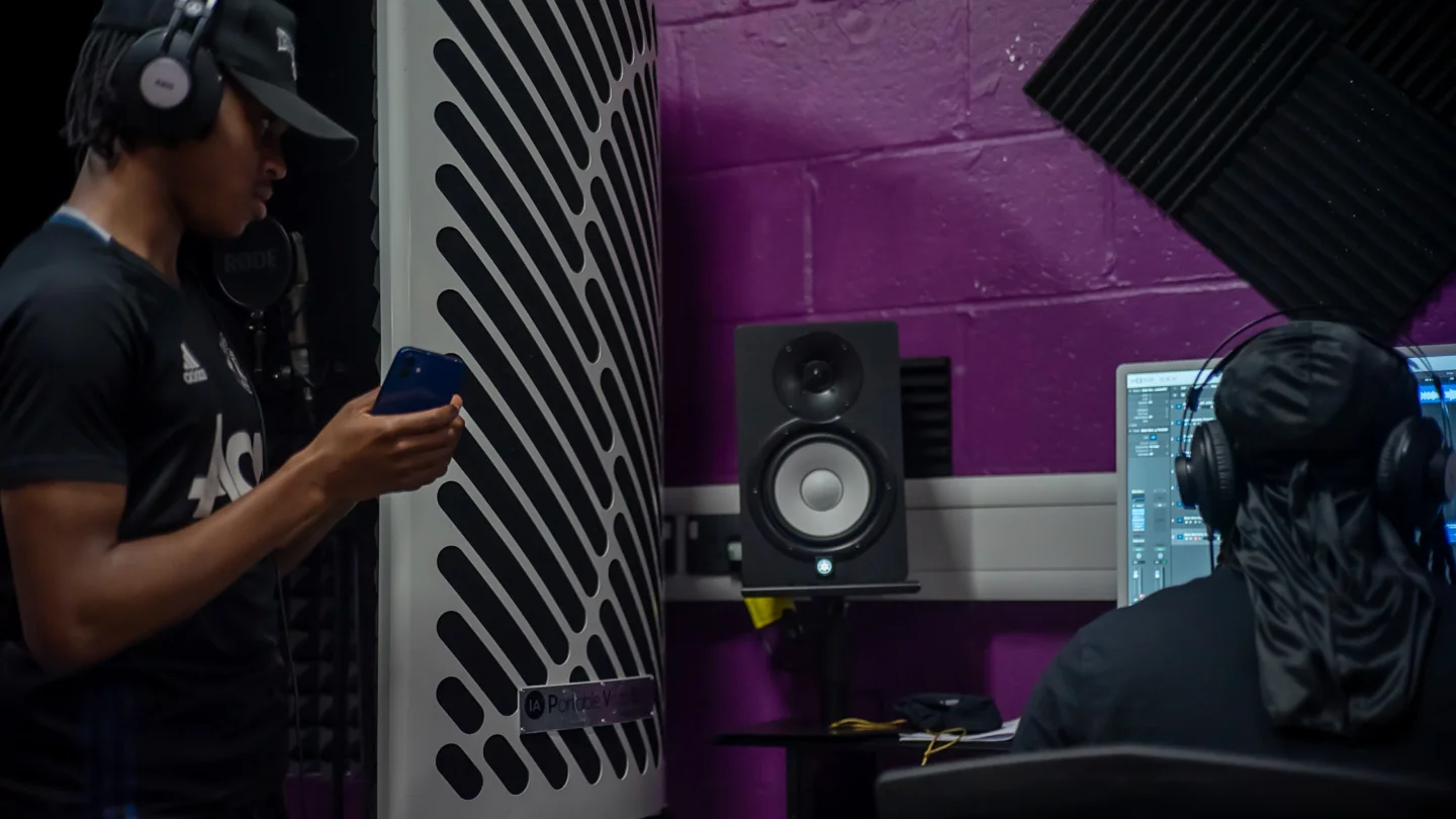 Two young men wearing headphones. One is facing a computer and speaker set up. The other is holding a microphone to his mouth and looking at his phone. Both look very engaged in what they are doing. The atmosphere is collaborative and creative.