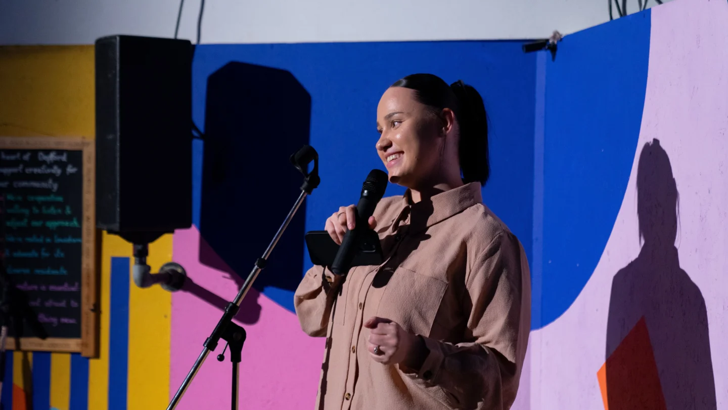 A young woman with a ponytail is holding a microphone in her hand and smiling brightly. She is being lit up by a spotlight in front of a colourful, vibrant indoor wall.