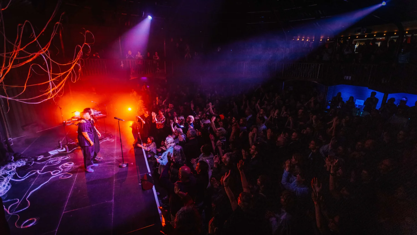 A photo of Kae Tempest on the Albany stage. They are looking out from the stage into a busy audience. They are lit with red and purple lighting and strings are hanging up behind them.
