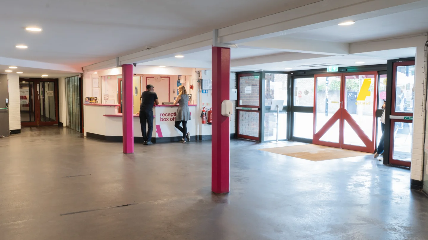 A photo of the Albany foyer, looking out towards the reception desk and front doors. There are two people stood chatting at reception and a couple of pink pillars in the middle of the space.
