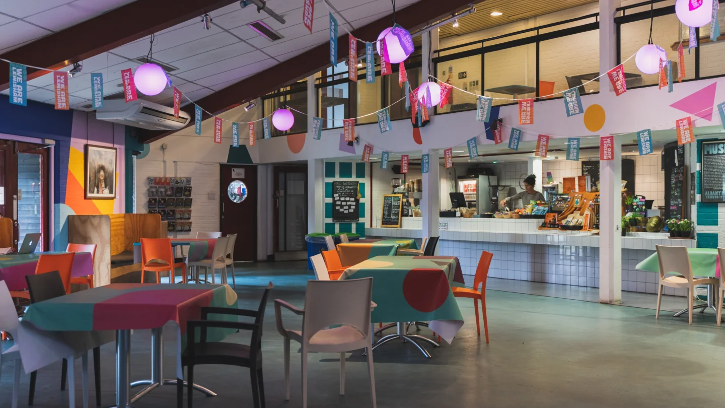 A photo of the Albany cafe. There are empty tables and chairs, and behind them is a woman with curly hair stood behind the counter. There are colourful tablecloths on the tables and bunting hanging from the ceiling.