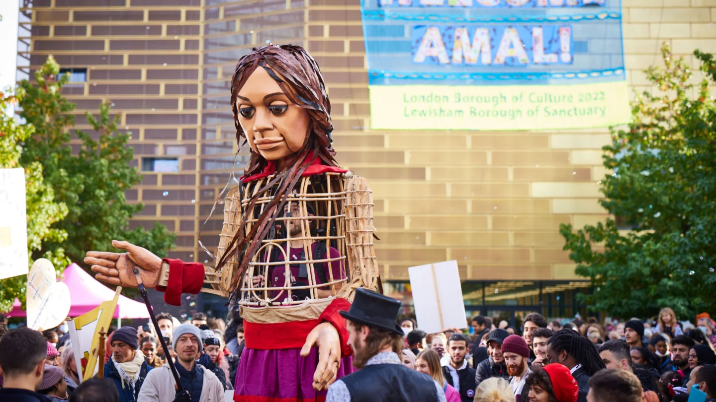 A photograph of a giant puppet of a young girl, with long brown hair and wearing a red, pink and gold dress. She is surrounded by a crowd, in front of a gold building (Deptford Lounge) with a blue banner hanging from it that says 'Welcome Amal'.
