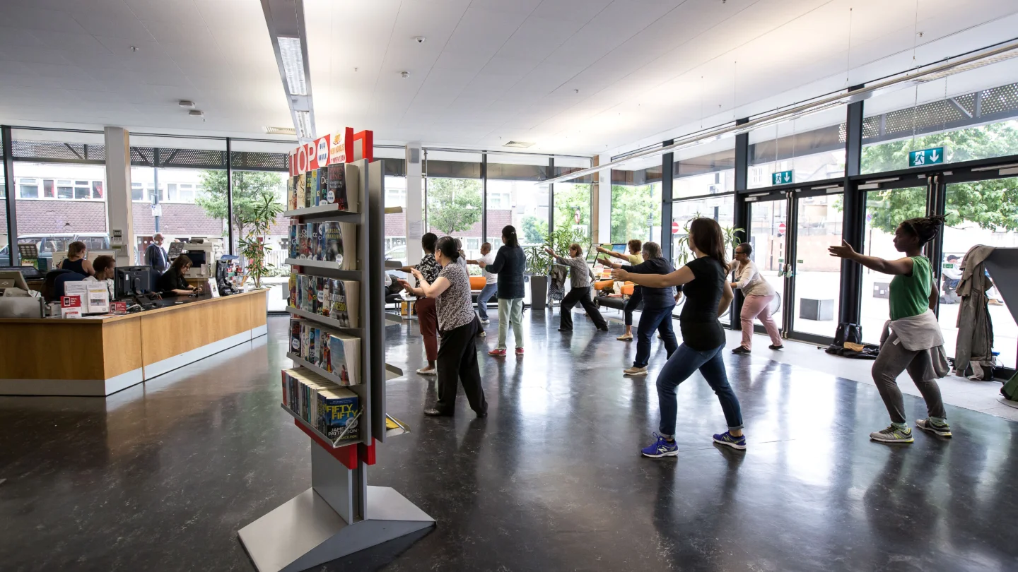 A photograph of a group of people doing Tai Chi in the foyer of Deptford Lounge. They are all standing, facing the same direction, with their legs apart and arms outstretched.
