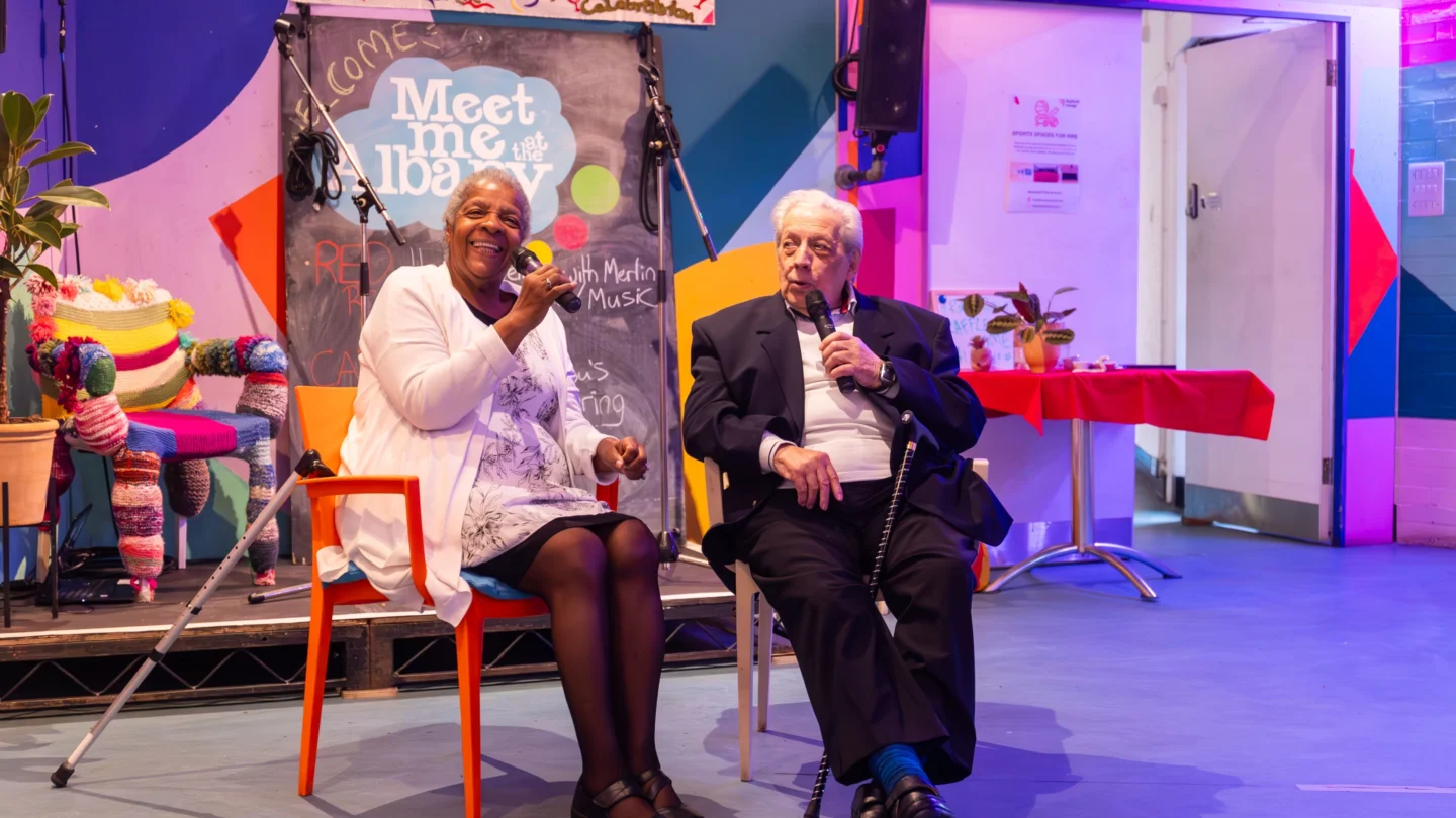 Elderly woman and man smiling, seated with microphones, against a colorful stage backdrop. Vibrant, inviting atmosphere with warm lighting.