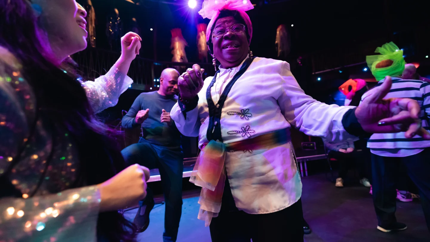 A person, dressed up in scarves, dances happily with others, creating a festive atmosphere filled with joy and movement. They are in a dark room with colourful lights and further people in the fore and background are similarly dressed up and wearing sparkly clothes.
