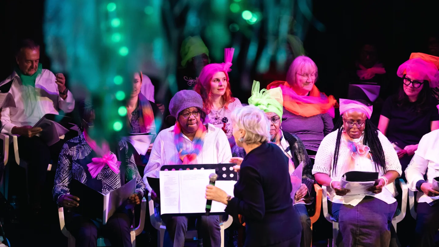 A vibrant choir lit by pink stage lights, featuring singers of various ages, performs under the guidance of an enthusiastic conductor.