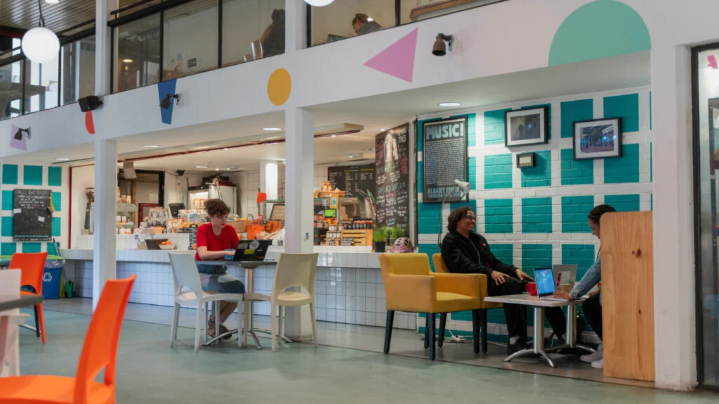 A photo of the Albany cafe. The walls are coloured by a bright mural. One woman in a red top is sat at a table working on a laptop, and at another table a man and a woman sit having a conversation.