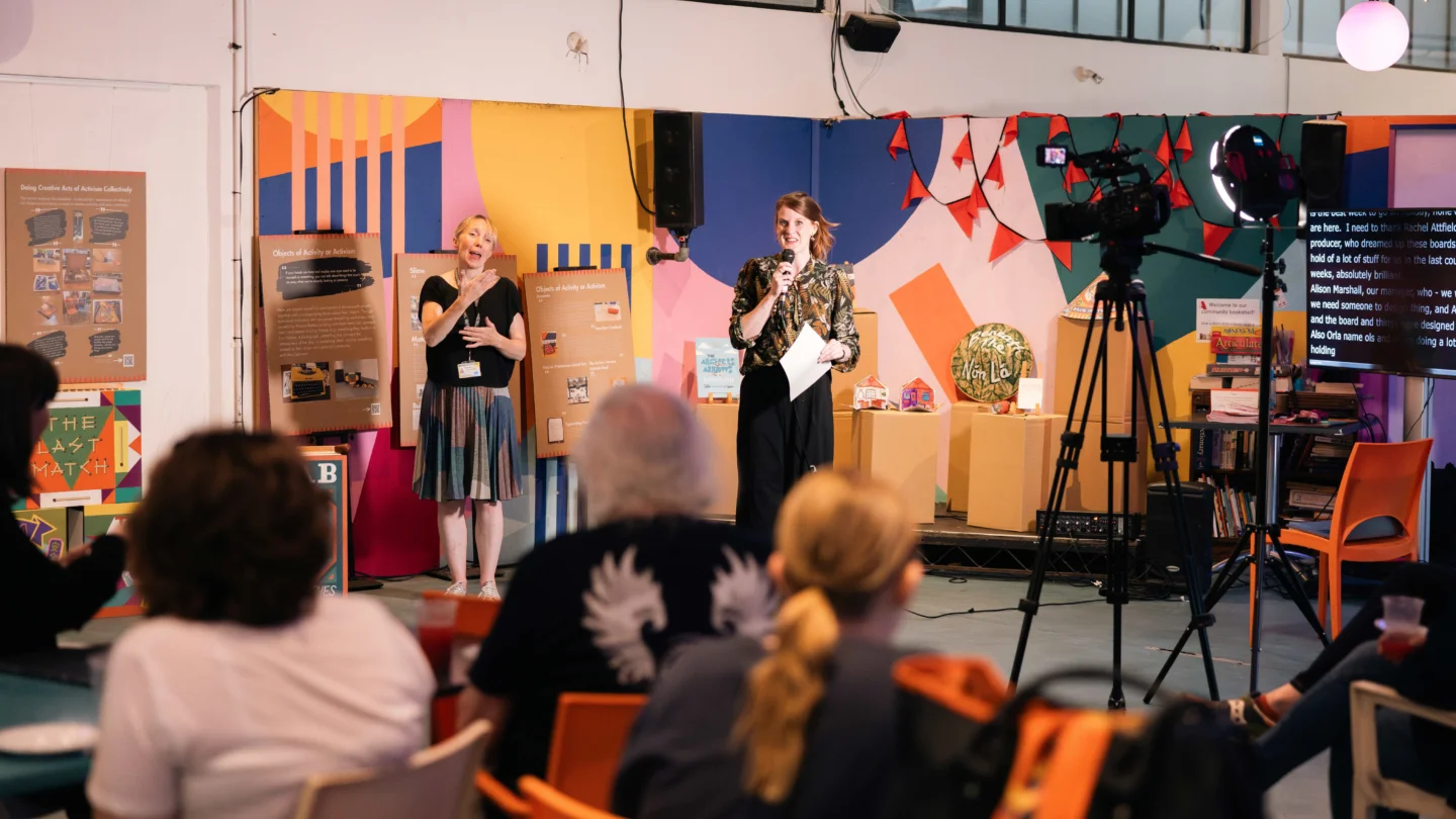 A photo of an event in the Albany cafe. Two white women are stood on a small stage in front of a brightly coloured mural, speaking in to microphones. An audience is sat watching them and there is also a camera filming them.