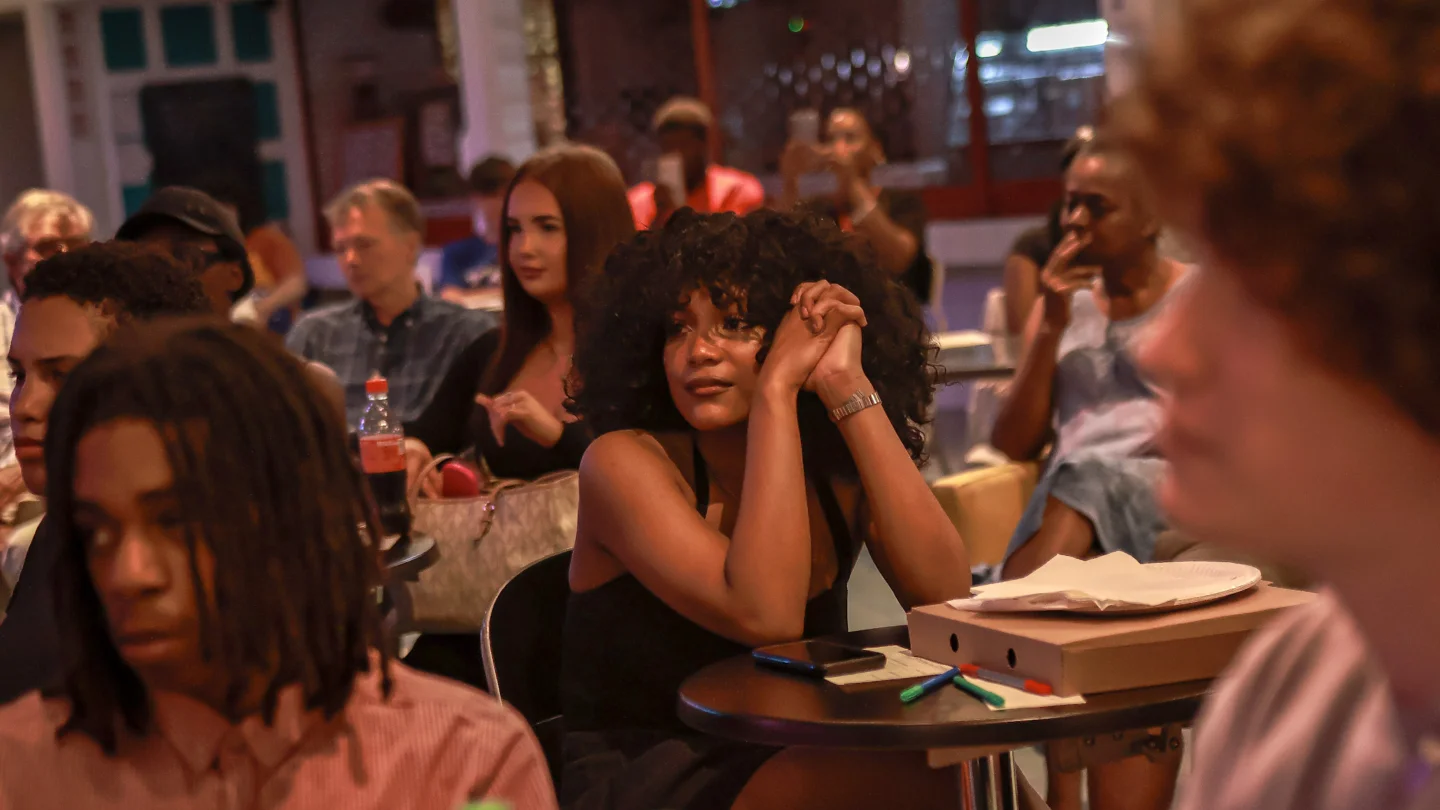 A photo of a group of young people sat at tables in the Albany cafe watching a performance. In the centre of the image is a woman with an afro wearing a black top. She is gazing intently at the performance, which is out of shot.