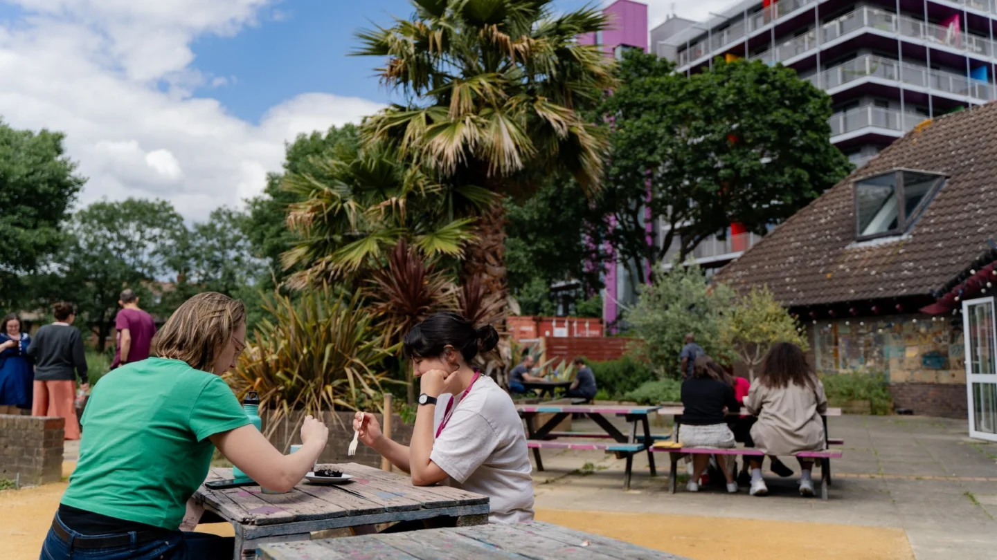 A photo of two women sat at a picnic table in the Albany garden eating cake. In the background are more people sat at tables, a palm tree and other plants.