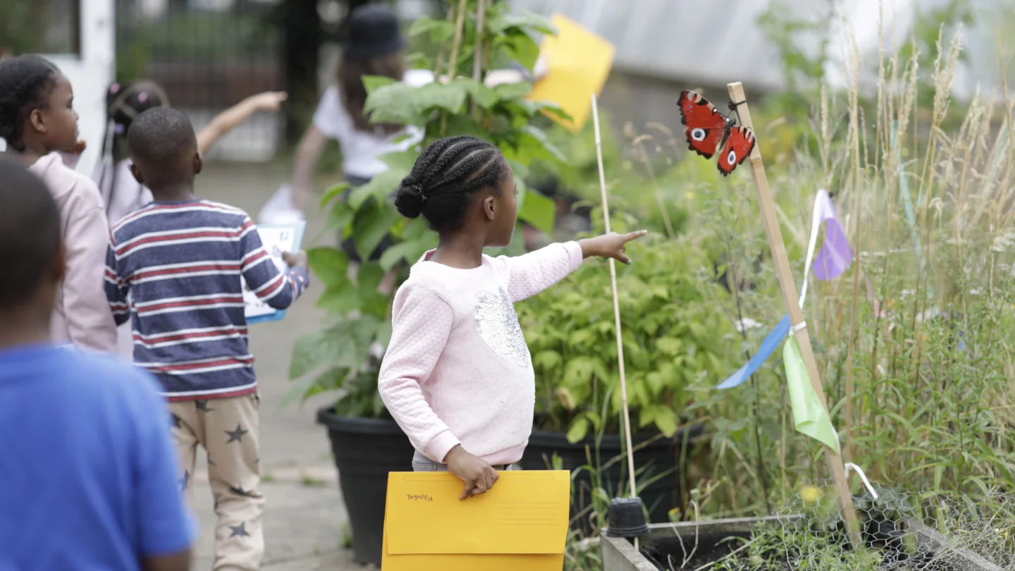 A photo of a young black girl with short braids pointing at a butterfly in the Albany garden.