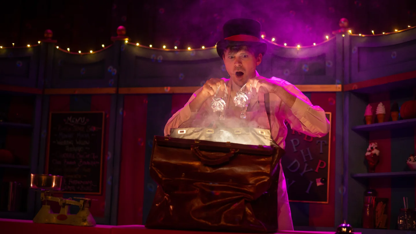 A photo of a performer on stage. The performer is a white man wearing a stripy shirt and a top hat. A light is glowing from a bag in front of him and he gazes at it, amazing. There is a string of fairy lights and a purple wash lighting behind him.
