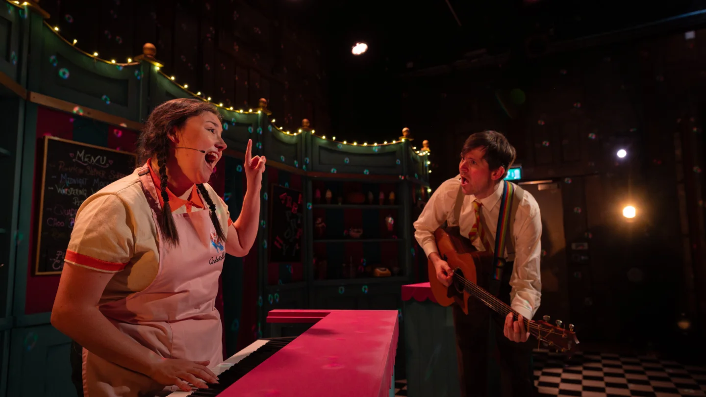Two performers sing a song on stage. On the left hand side, a white woman with plaits and wearing a pink apron plays a red piano. To her right is a white man wearing a stripy suit and playing the guitar. There is a string of fairy lights behind them.