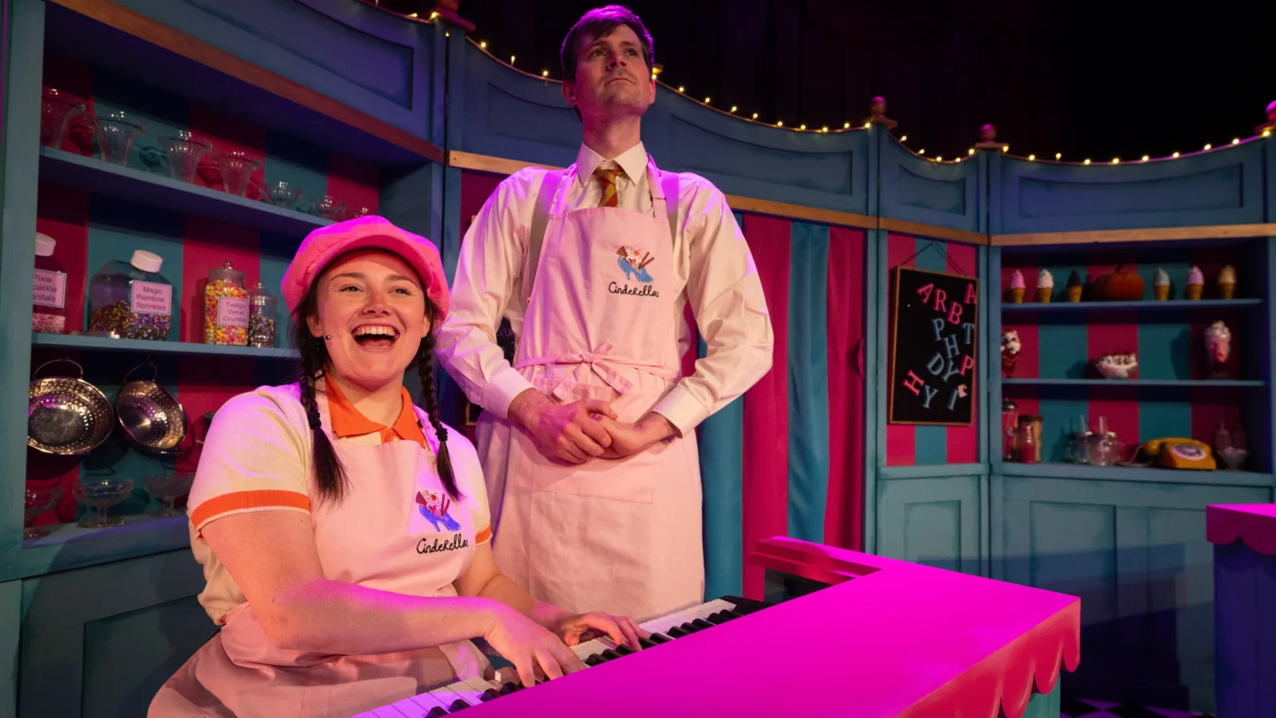 Two performers sing a song on stage. A white woman with plaits and wearing a pink hat and pink apron sits behind a piano, playing the piano and singing. Behind her a white man, also wearing a pink apron, stands formally behind her with his hands clasped in front of him. Behind them are shelves stocked with jars, as if they are in a shop.