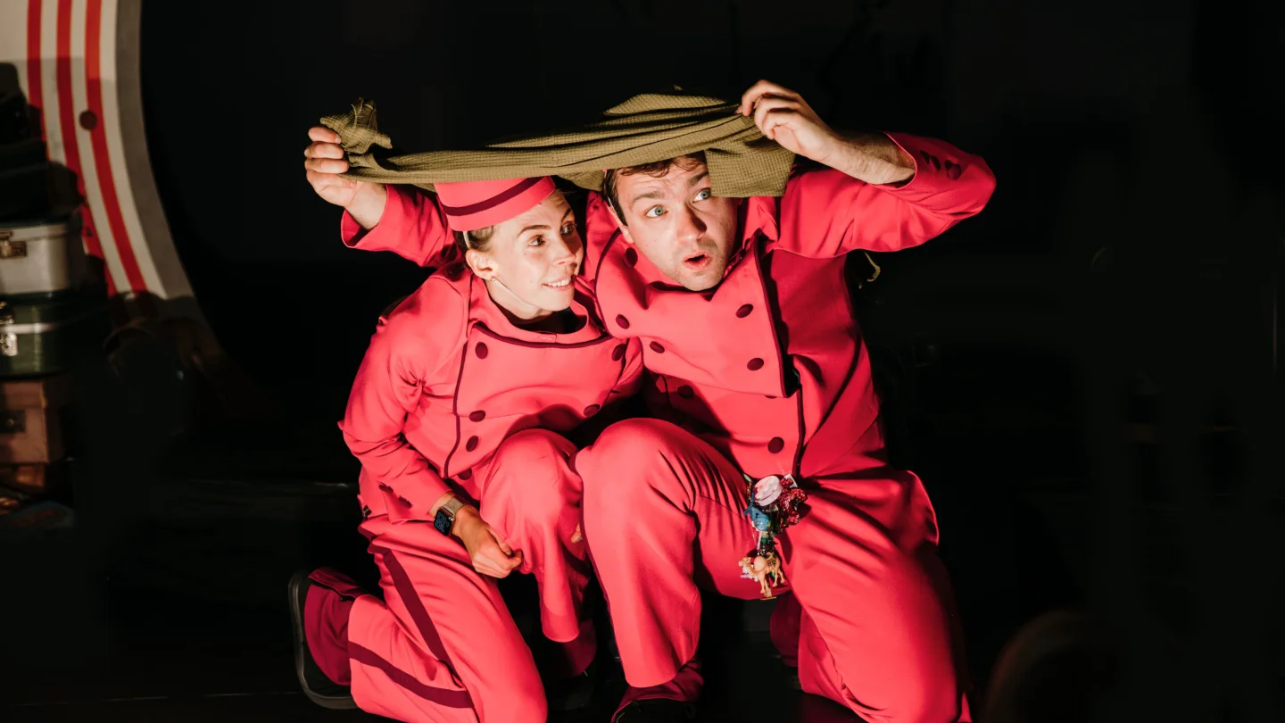 A photo of two performers wearing run suits, crouched down under a blanket.