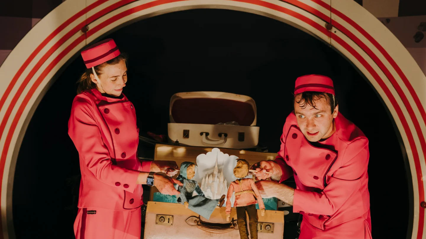 Two performers wearing red suits and hats stand on either side of an open suitcase. They are each holding a puppet and behind them inside the suitcase is a pop-out landscape of a castle amongst the clouds.
