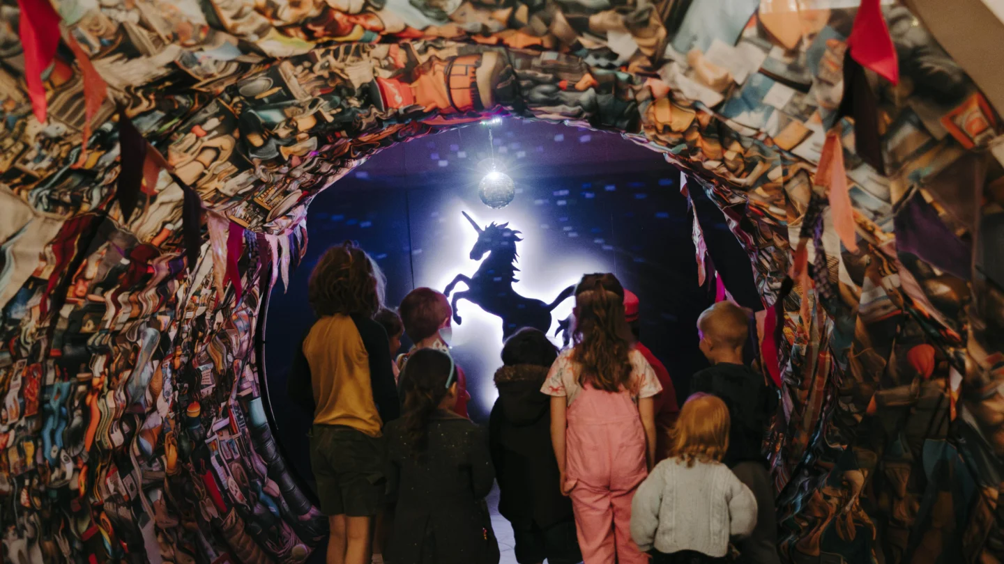 A photo of a group of children in a tunnel. Their backs are to the camera and they are gazing at an illuminated purple unicorn at the end of the tunnel.