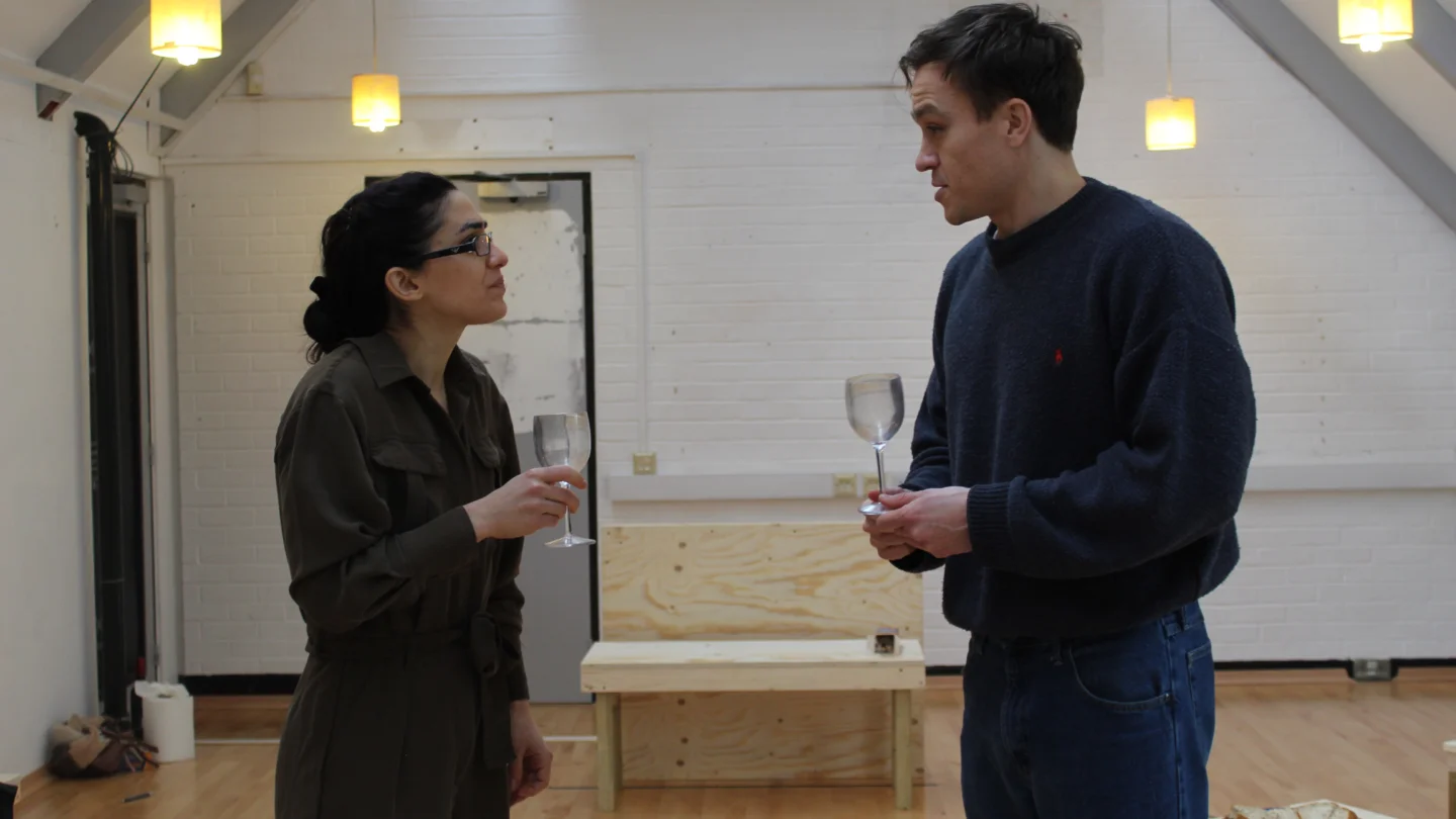 James Haddrell Cassie Bradley and Barney White in rehearsal for One Day When We Were Young. A white woman and white man stand face to dace holding empty wine glasses.