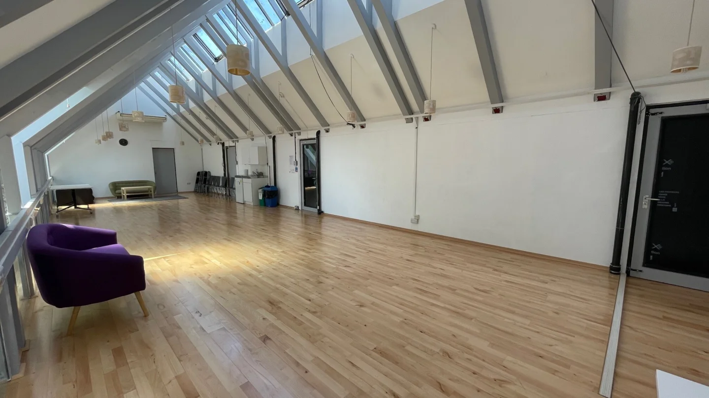 A photo of the Rehearsal Room at the Albany. It is an empty room with a wooden floor, white walls and grey exposed beams across the ceiling. Along the left wall is a purple sofa.