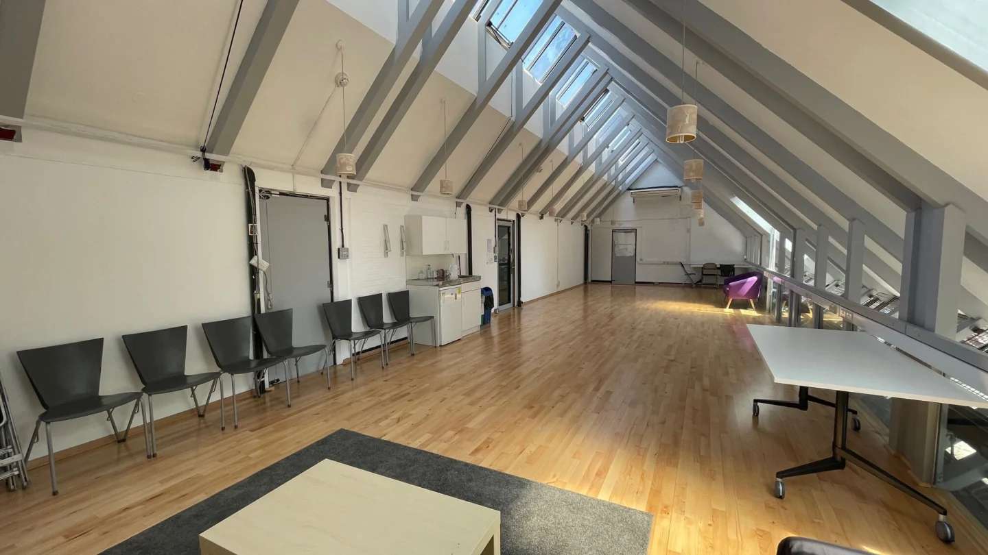 A photo of the empty rehearsal room at the Albany. It is a long room with a wooden floor, white walls and grey exposed beams on the ceiling. In the space are a row of chairs, a black rug, coffee table, white table and purple sofa.