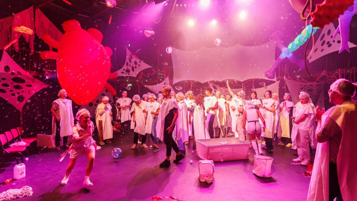 A lively, whimsical stage scene features people in white costumes and shower caps, brightly lit in pink. The backdrop is filled with colourful, abstract decorations.
