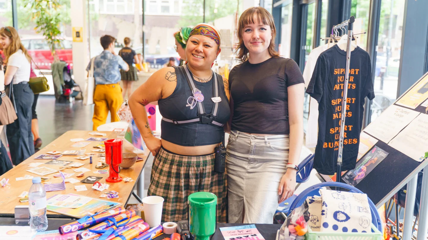 A photo of two white people with short hair stood behind a market stall with their arms around each other.