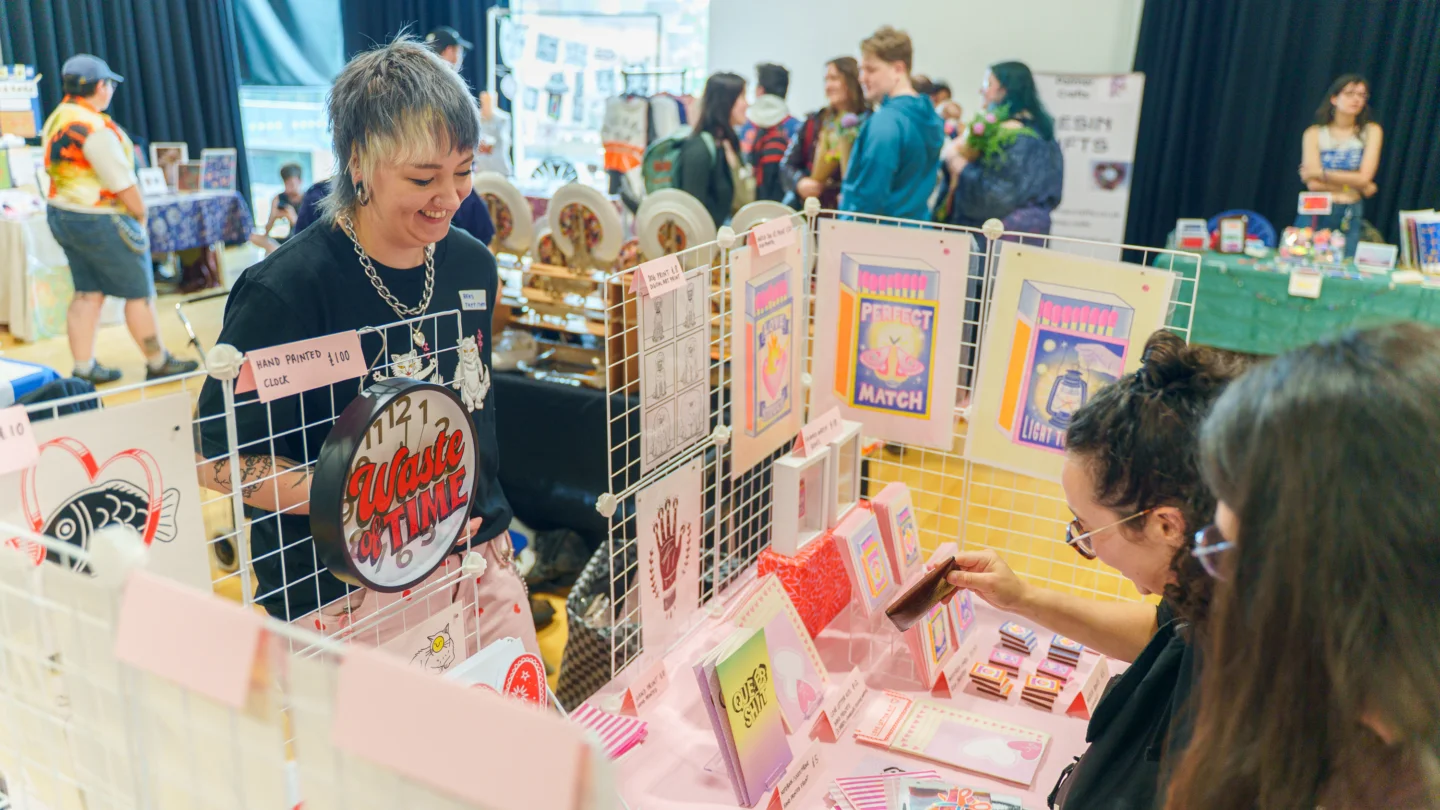 A photo of a person with short blonde hair peering over a craft stall selling stickers and cards.