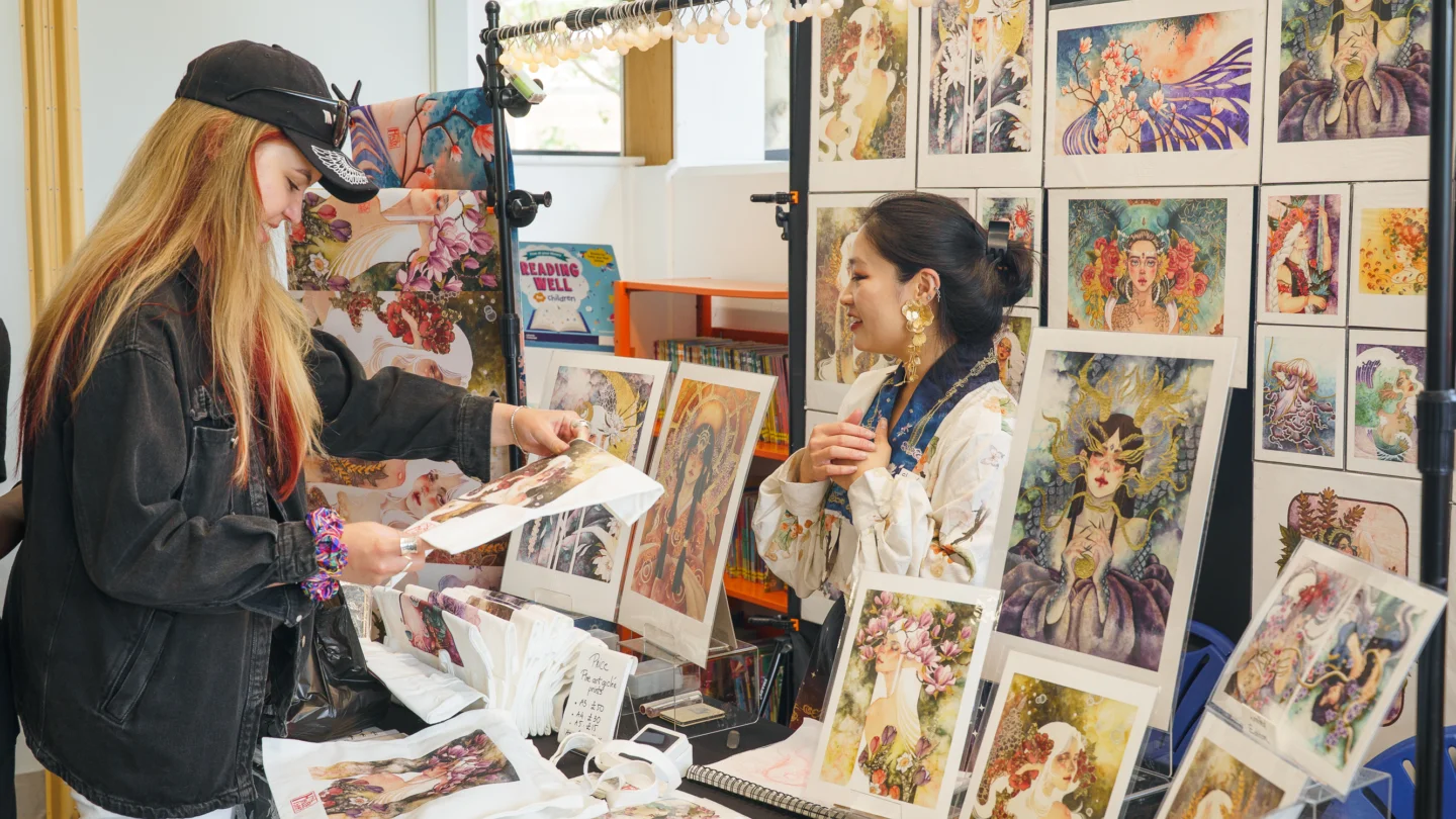 A photo of a person with long hair, wearing a black jacket and black cap, stood at a market stall full of colourful prints.