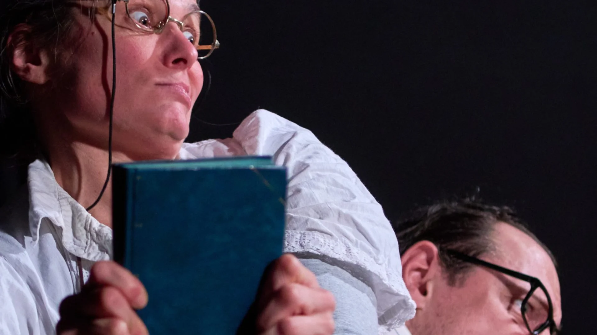 A photo of a woman wearing round glasses and a white shirt clutching a book. Behind her a man wearing glasses leans down - you can just see his head at the bottom of the photo.