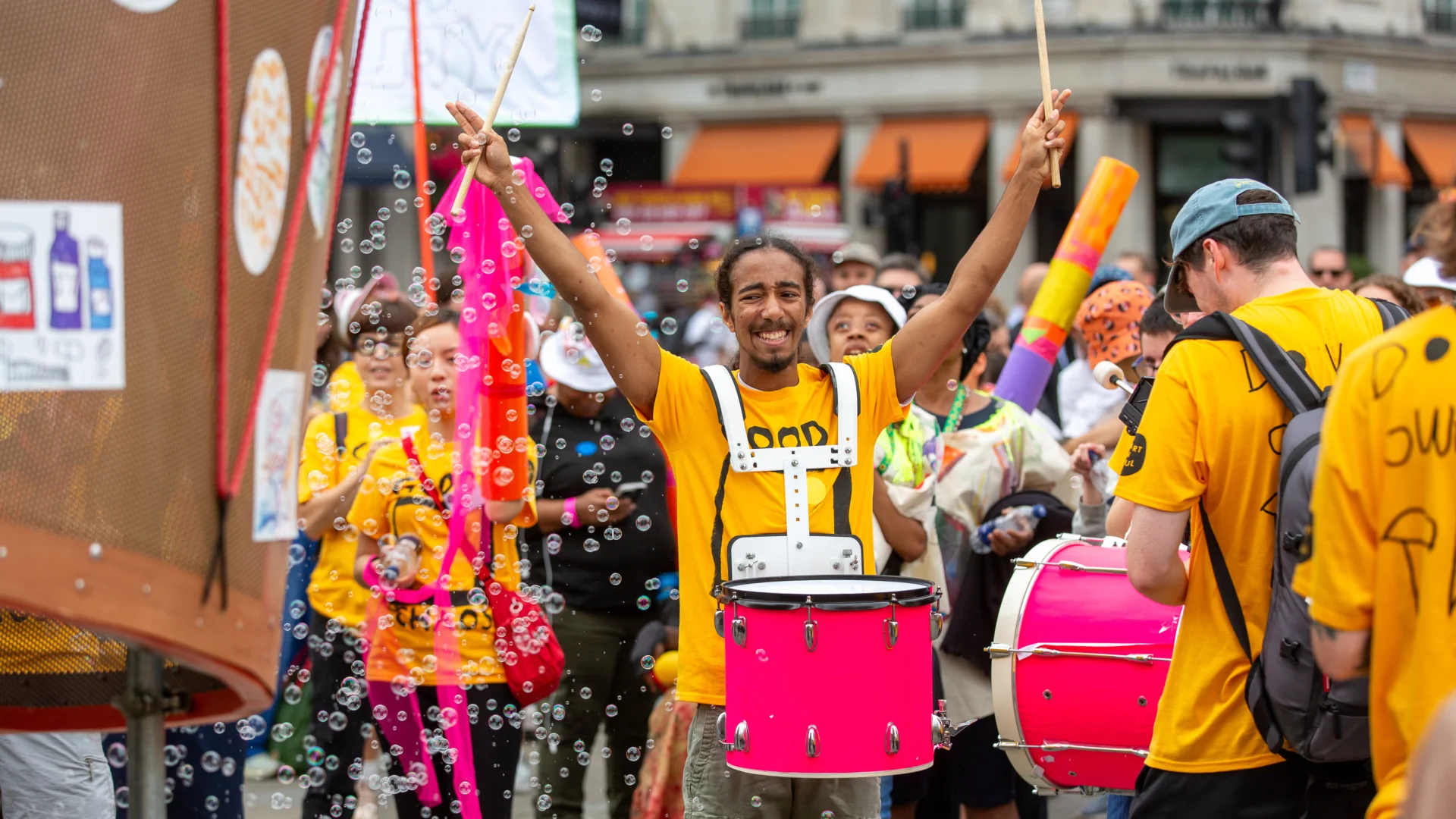 A photos of a band wearing yellow t-shirts on the street. A man in the centre of the tshirt is playing a pink drum and has his hands in the air