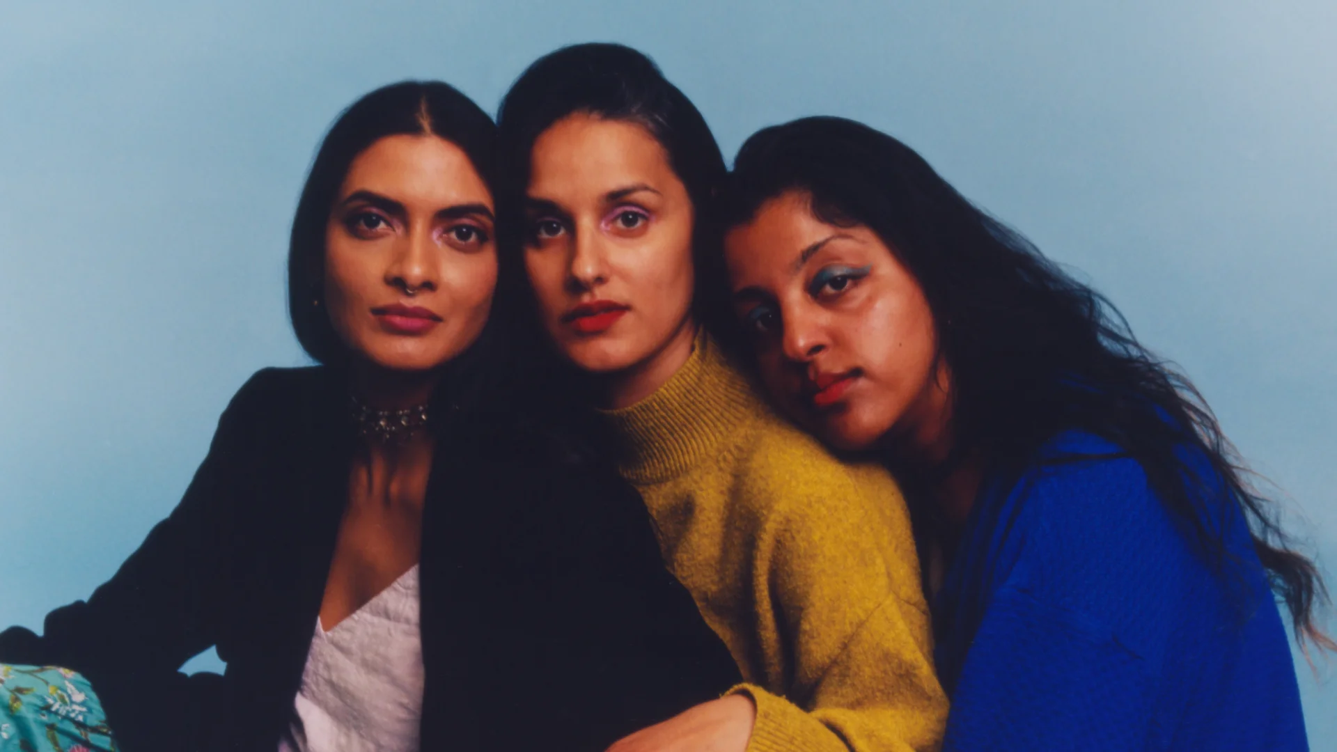 A posed photograph of three Asian women sat closely together and gazing into the theatre. They all have long dark hair and the backdrop behind them is pale blue.