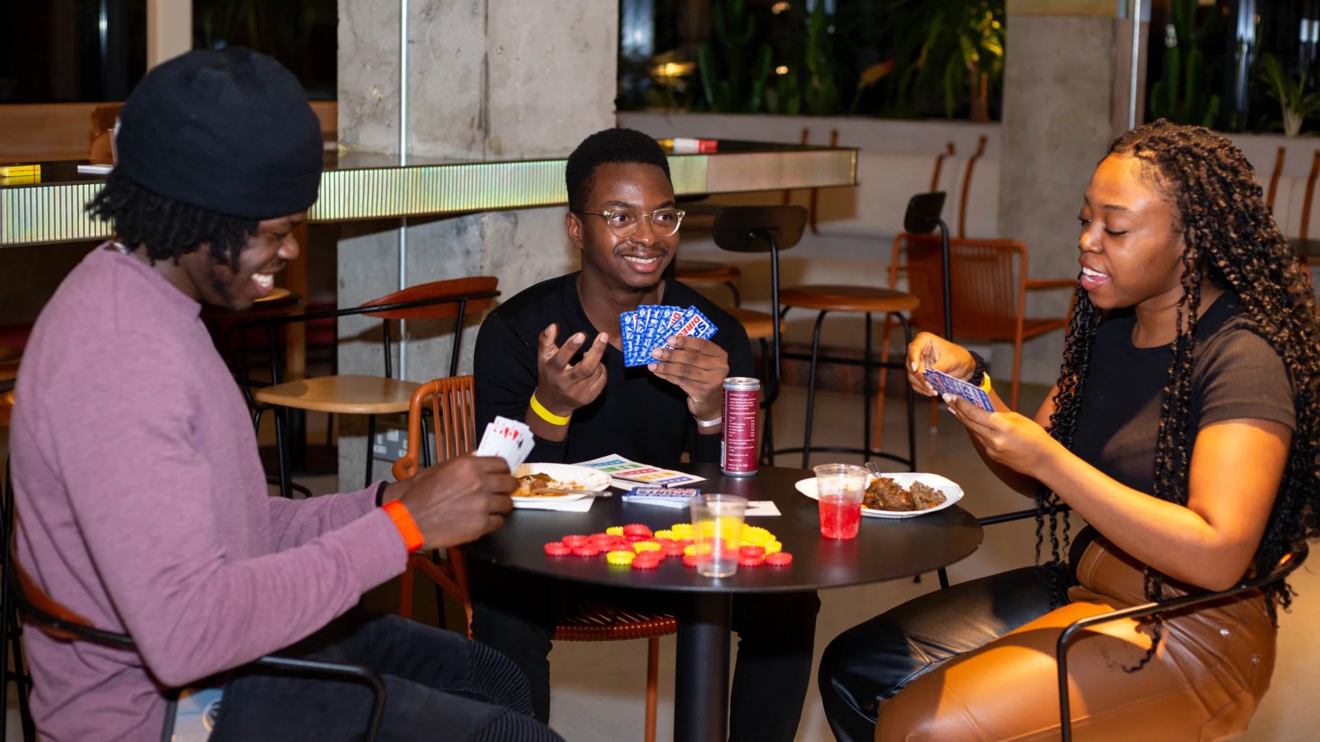 A photo of three people, two black men and one black woman, sat around a table playing cards and eating snacks.